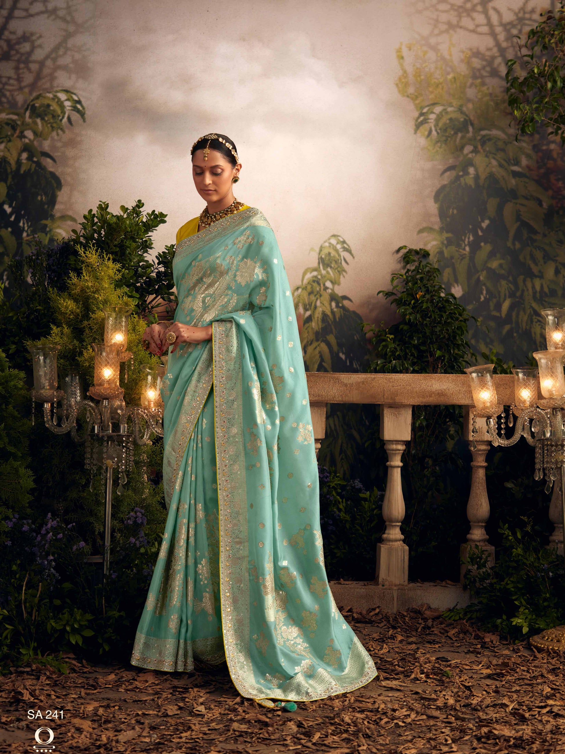 Woman in a light blue saree standing in an elegant outdoor setting with candles and greenery.