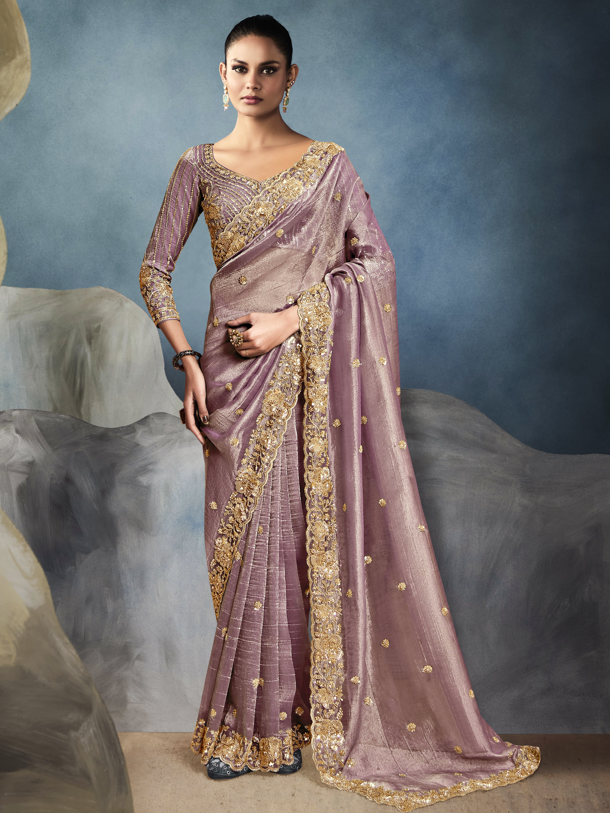 Woman wearing a mauve saree with gold embroidery against a textured gray background