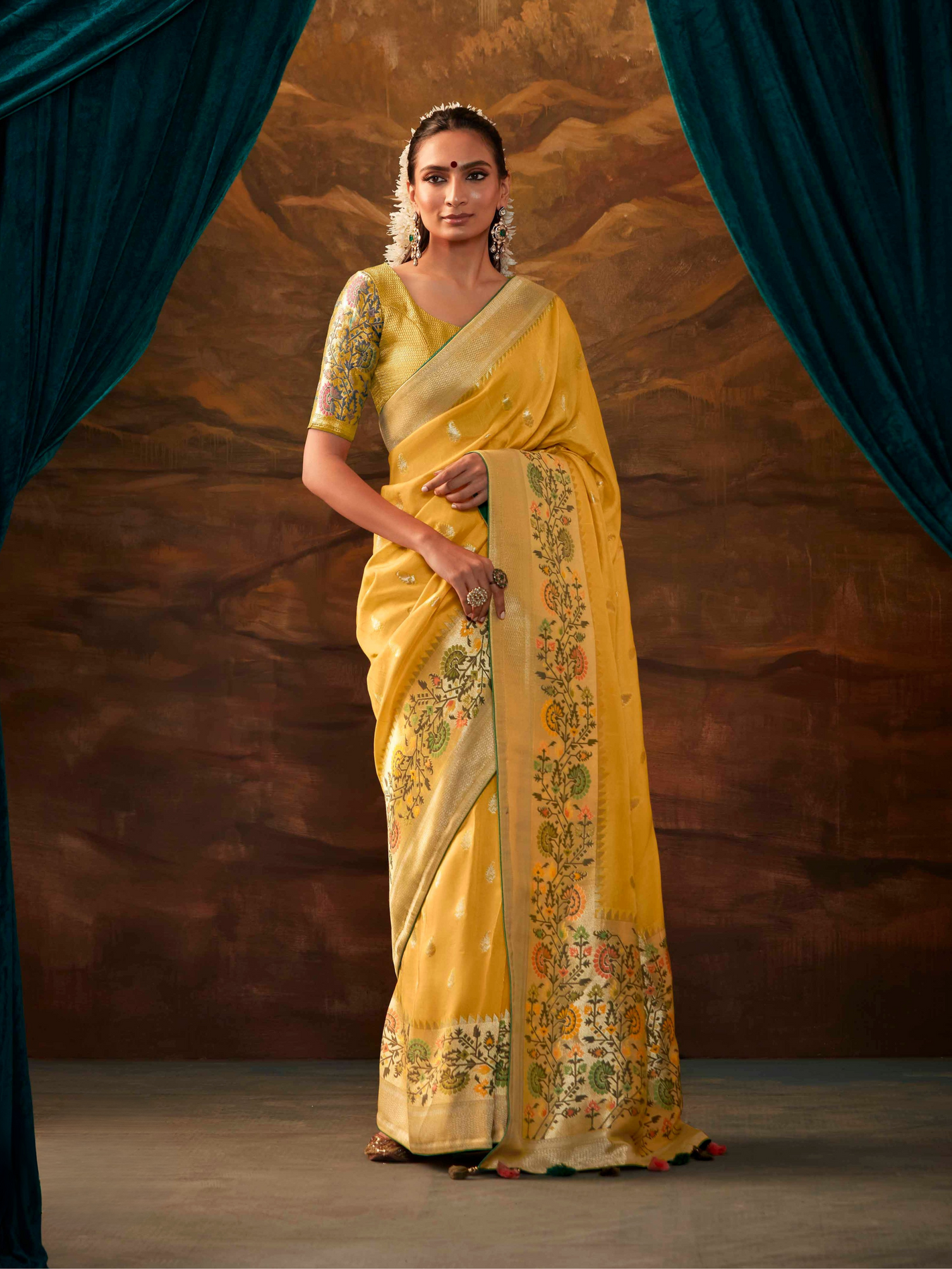 Woman wearing a yellow saree with floral patterns against a brown curtain backdrop
