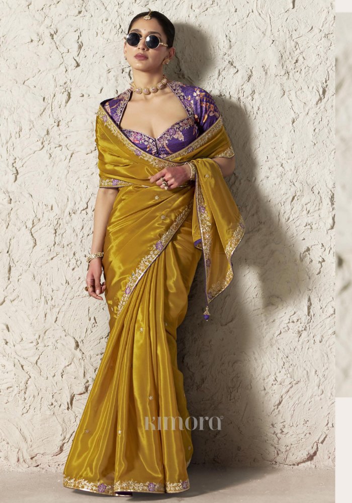 Woman wearing a yellow saree with a purple blouse against a textured white wall.