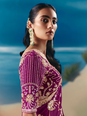 Woman wearing a purple saree with gold embroidery against a blurred natural background