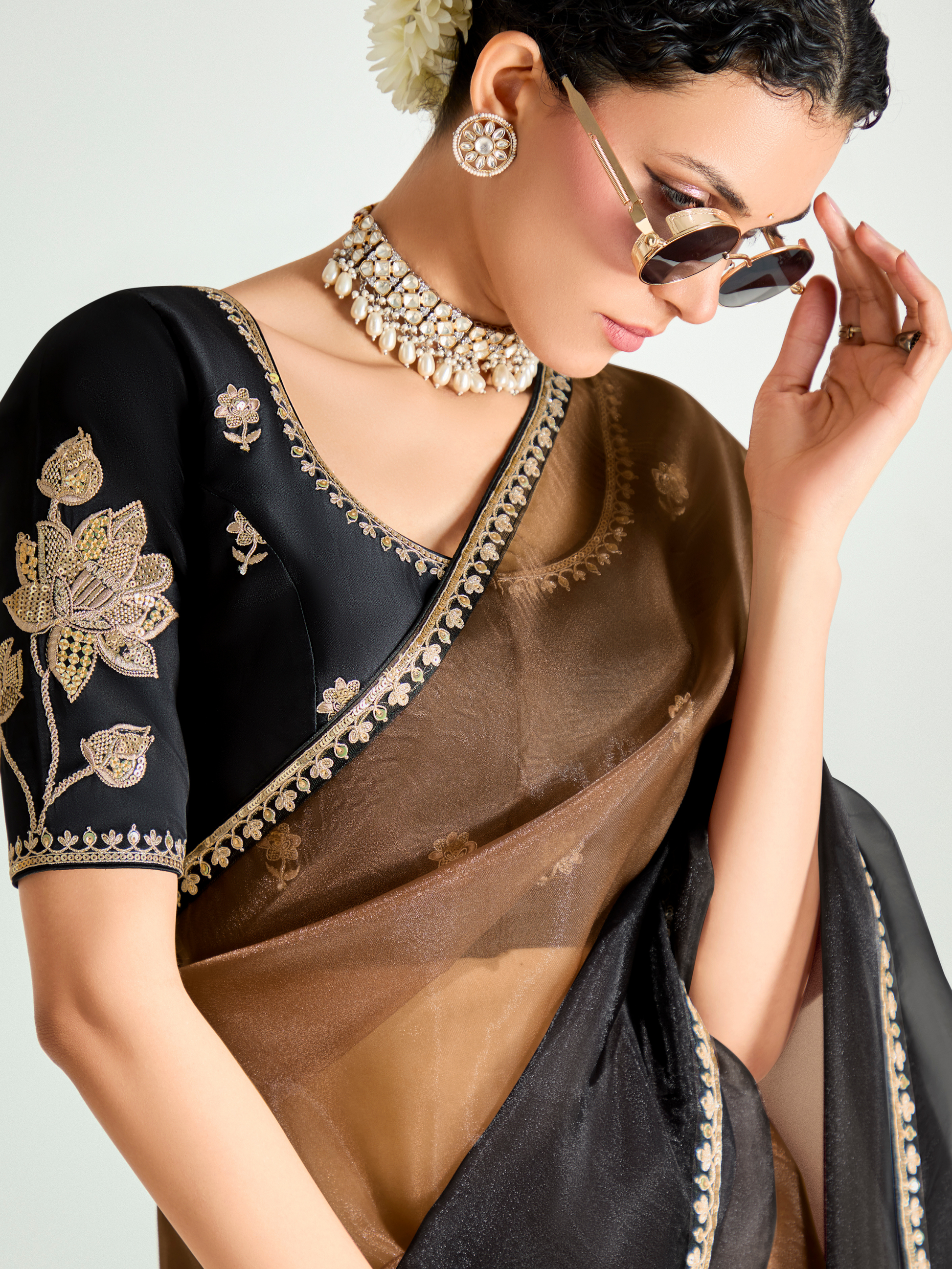 Woman wearing a black and gold saree with jewelry and sunglasses on a light gray background