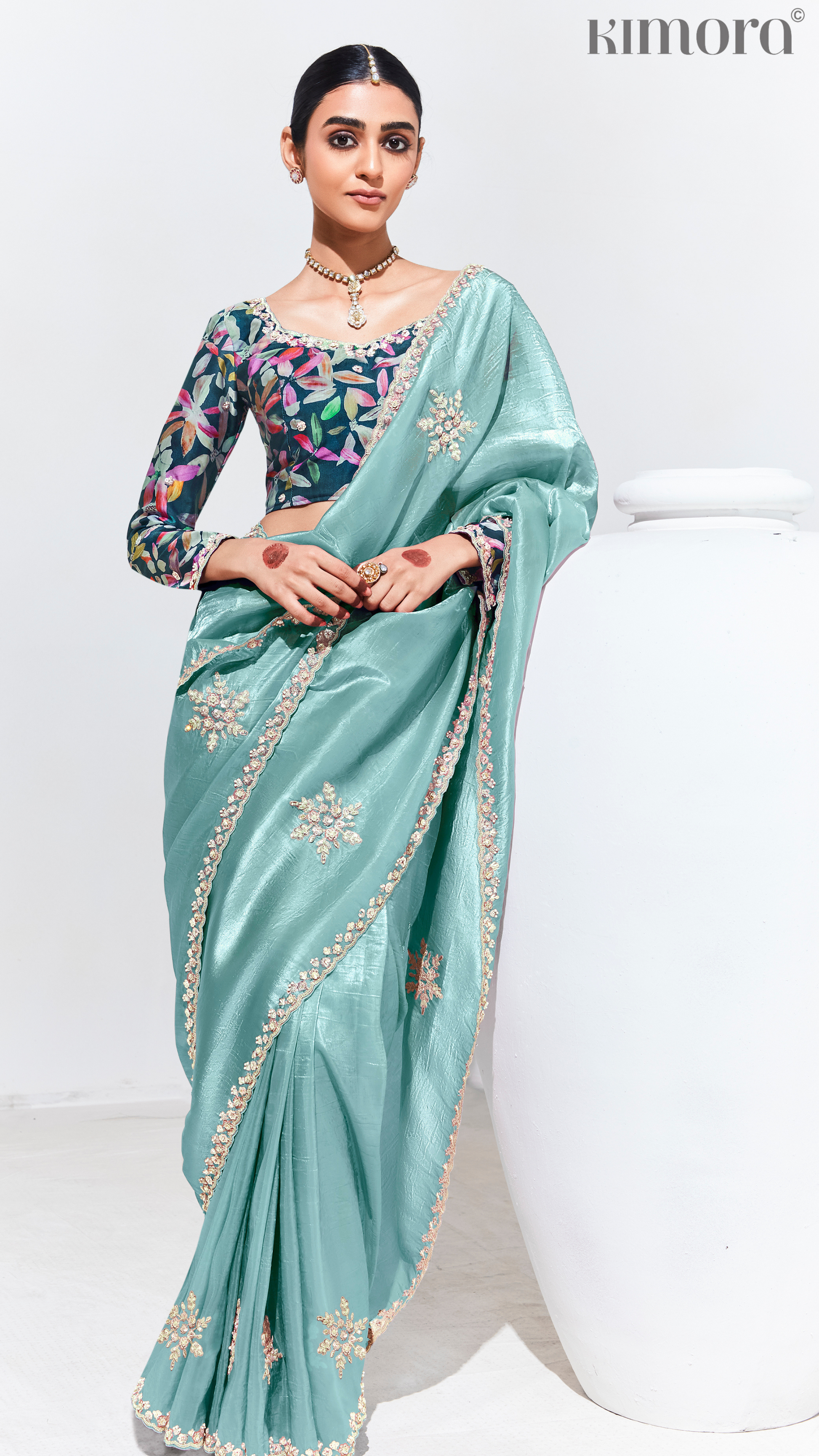 Woman wearing a light blue saree with floral blouse, sitting on a white chair against a white background.
