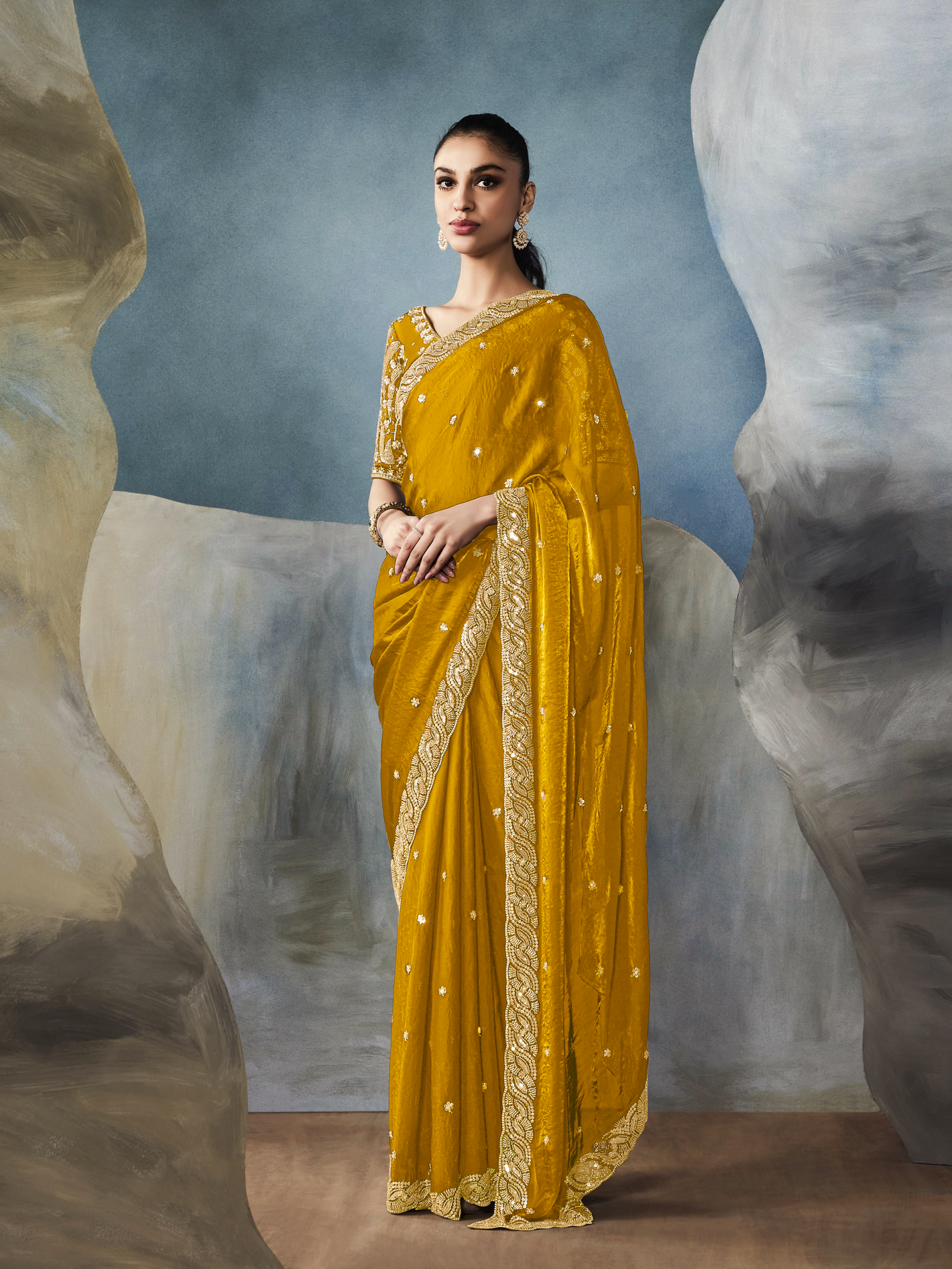 Woman wearing a yellow saree with gold border against a textured wall background