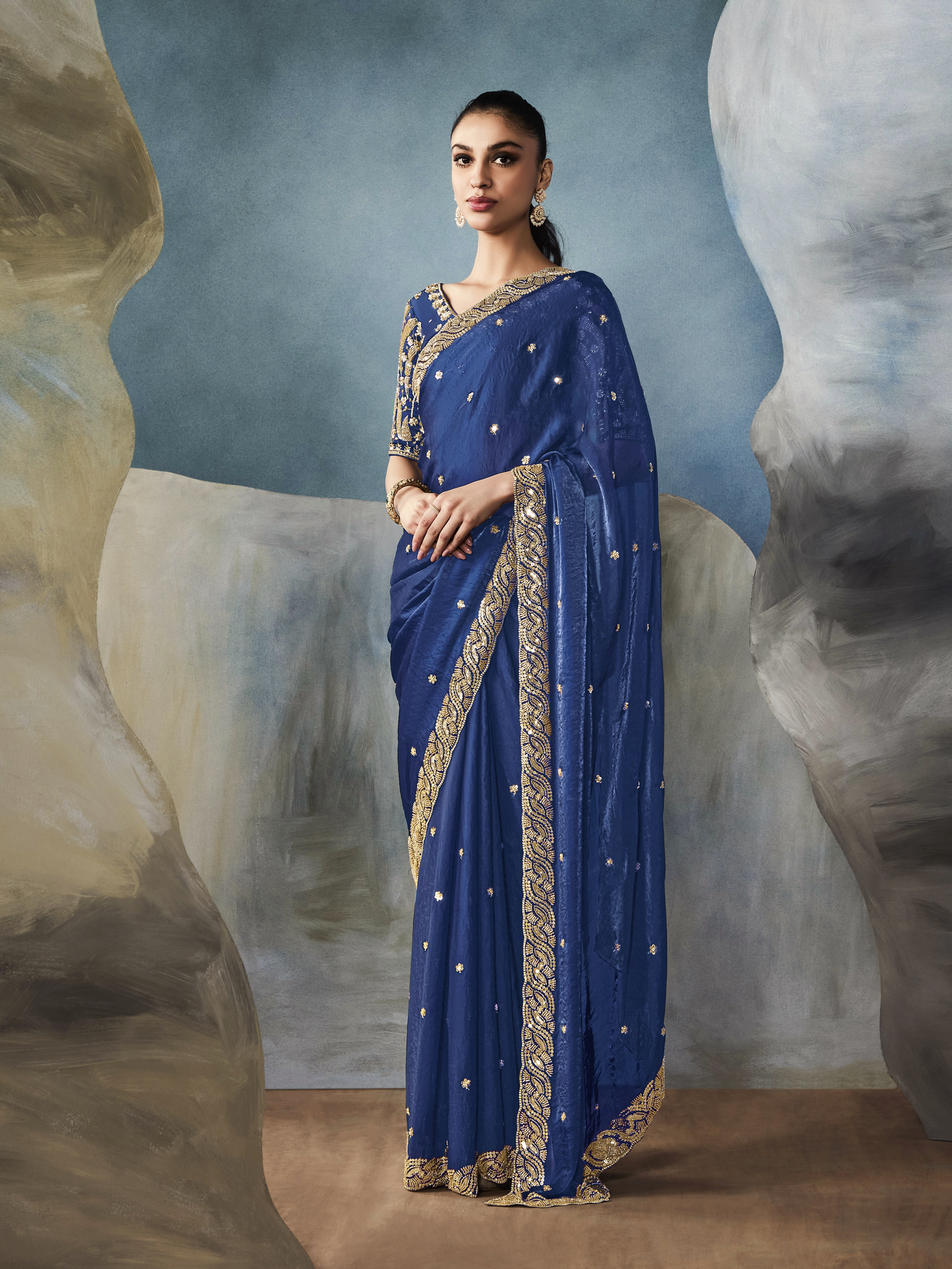 Woman wearing a blue saree with gold embroidery against a textured wall background