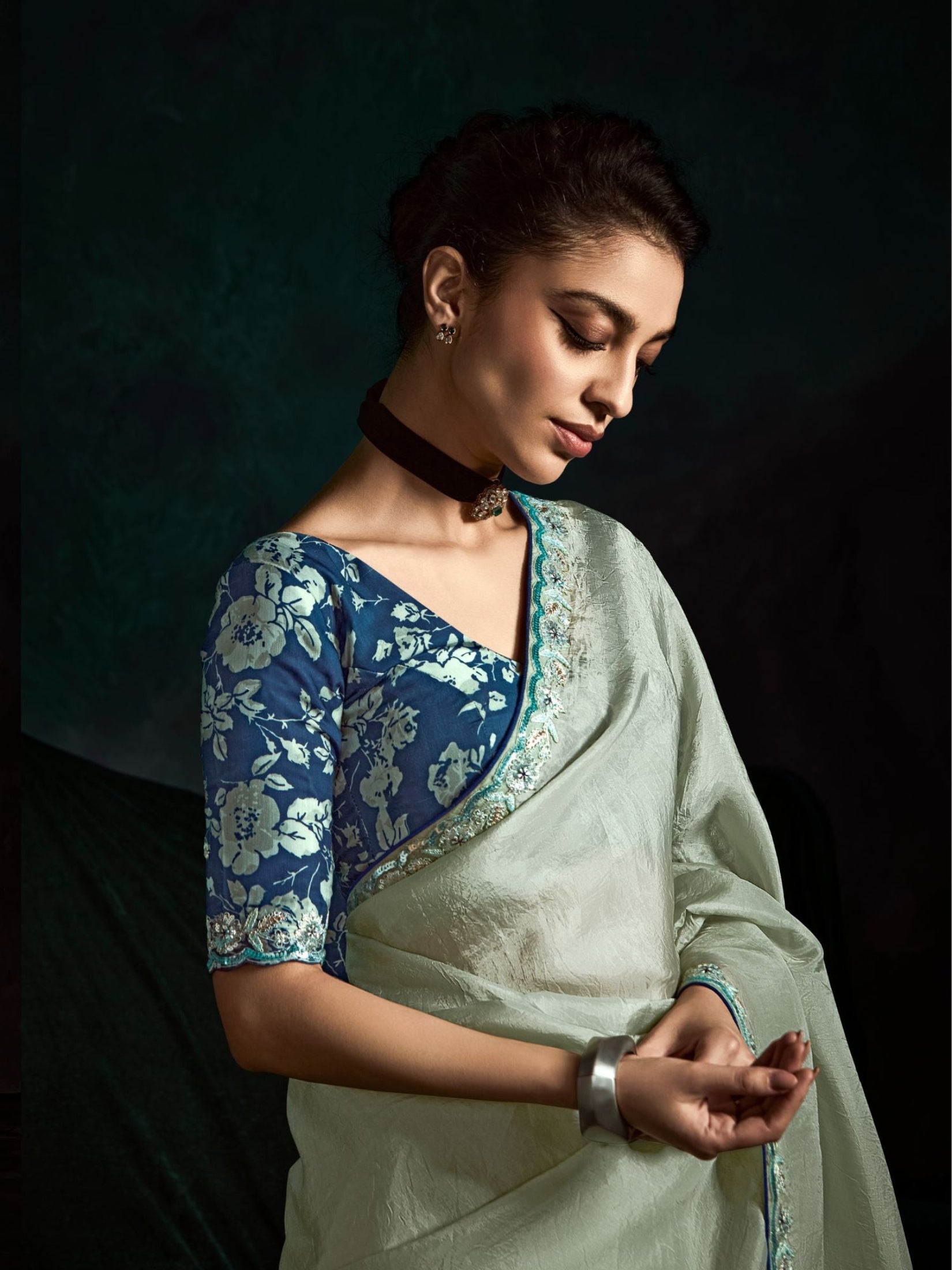 Woman wearing a blue floral blouse and light green saree against a dark background