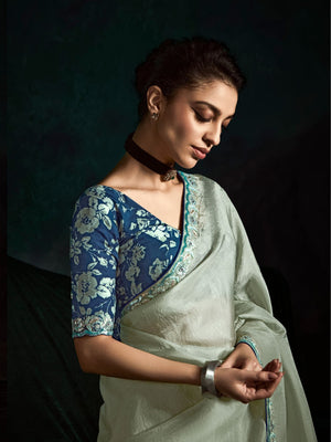 Woman wearing a blue floral blouse and light green saree against a dark background