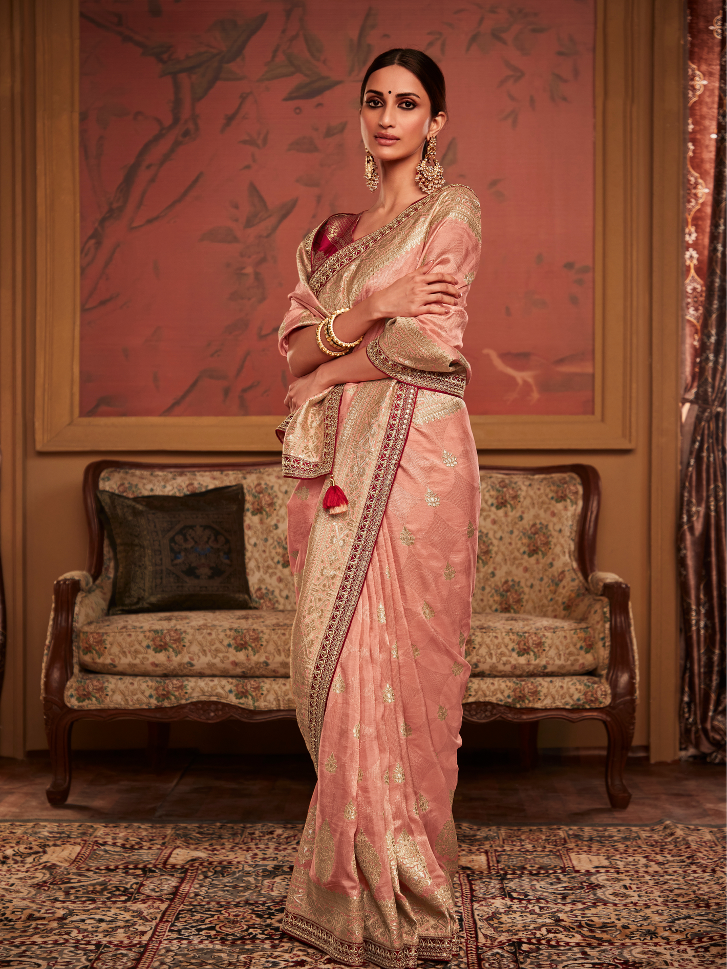 Woman in a traditional pink saree standing in an elegant room with a sofa and decorative wall.