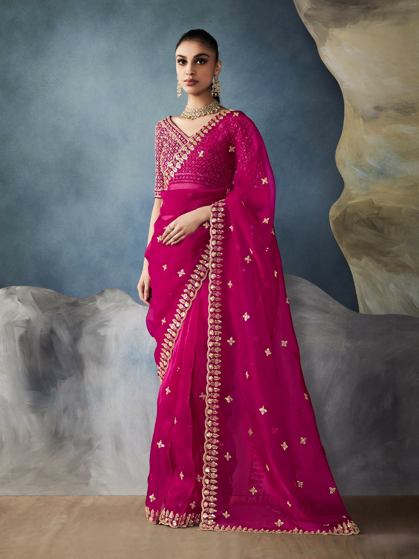 Woman wearing a vibrant pink saree with gold embroidery against a textured gray background