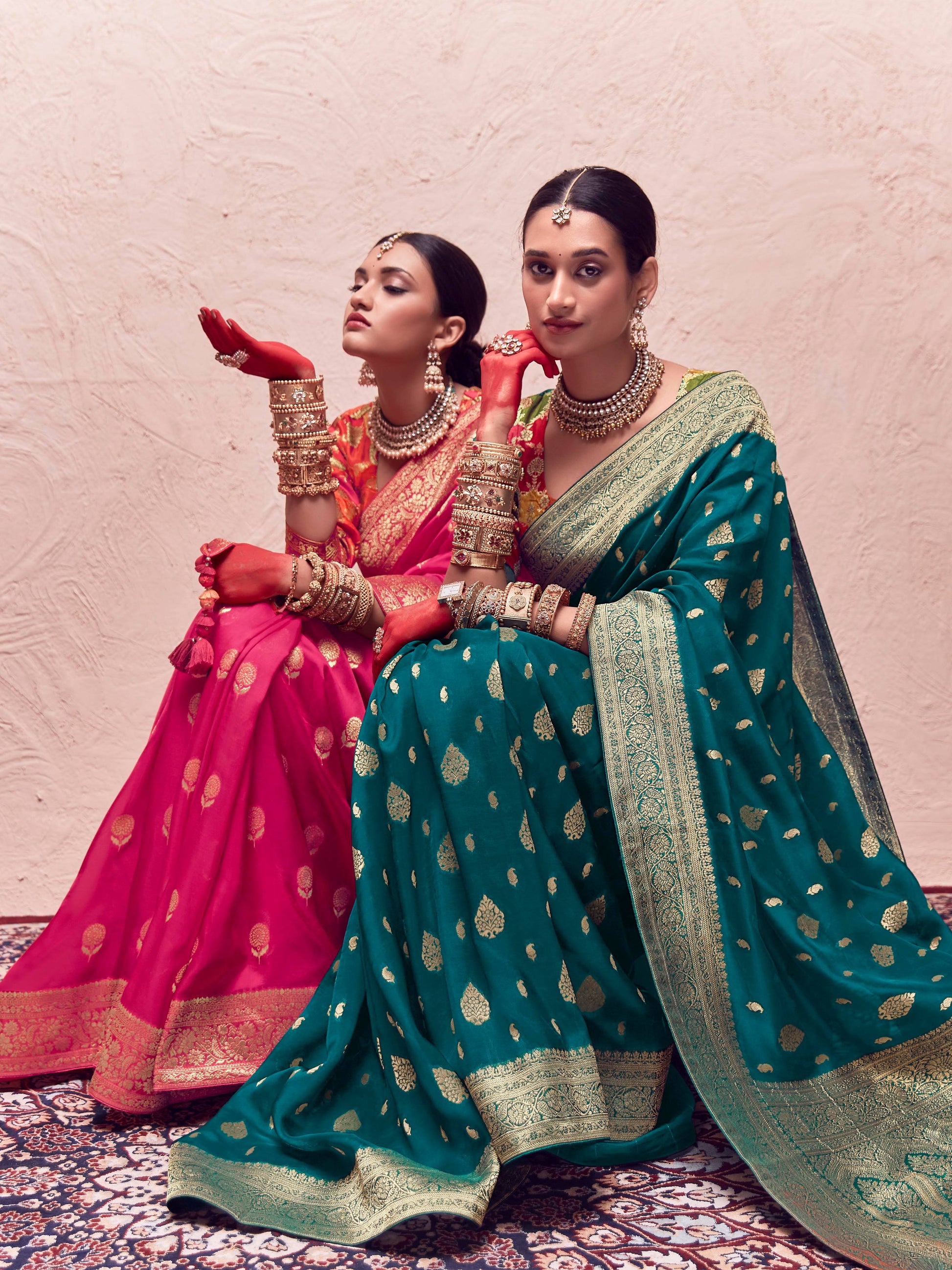 Two women in traditional sarees sitting on a patterned rug.