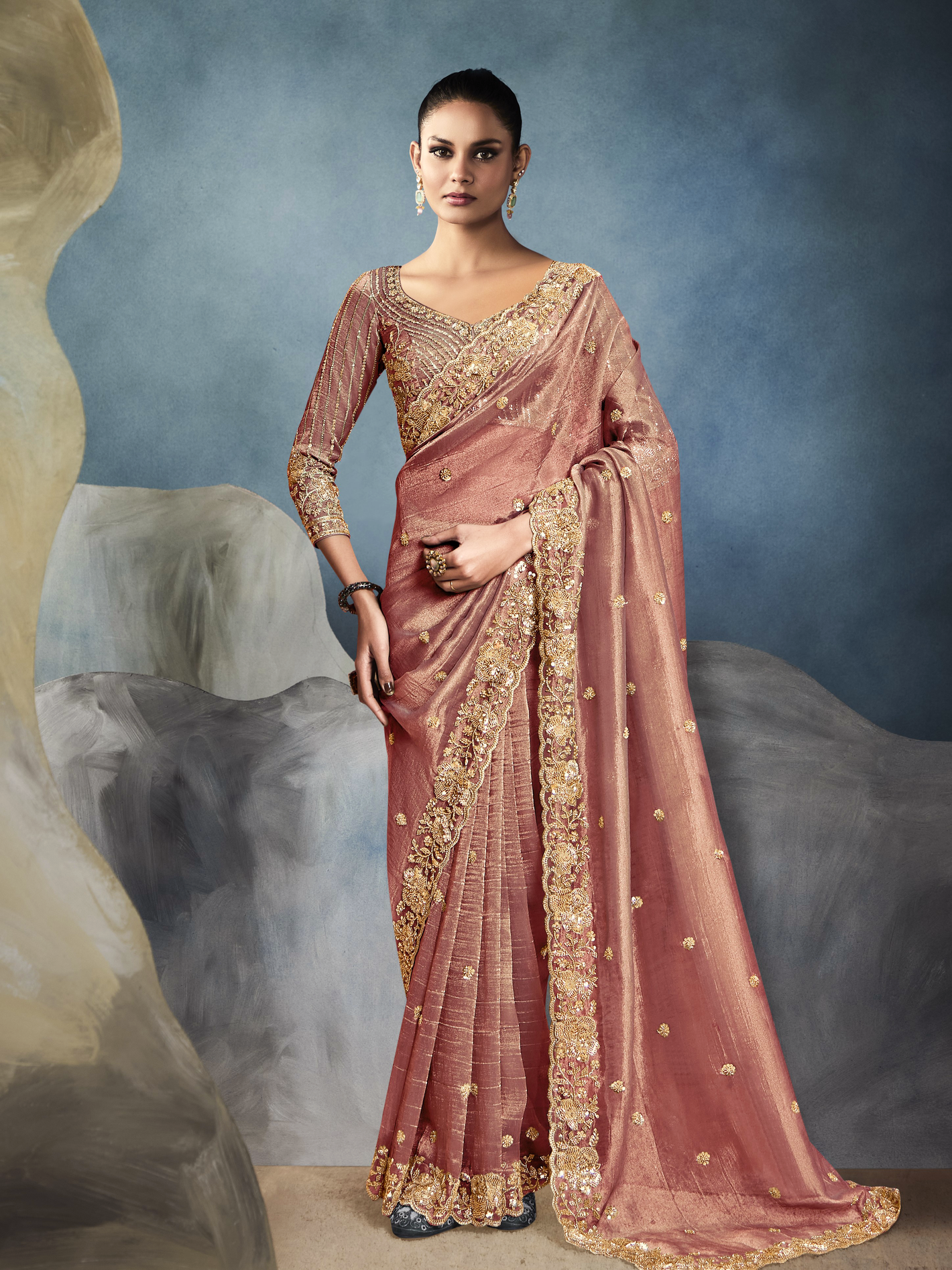Woman wearing a traditional pink saree with gold embroidery against a textured blue and gray background