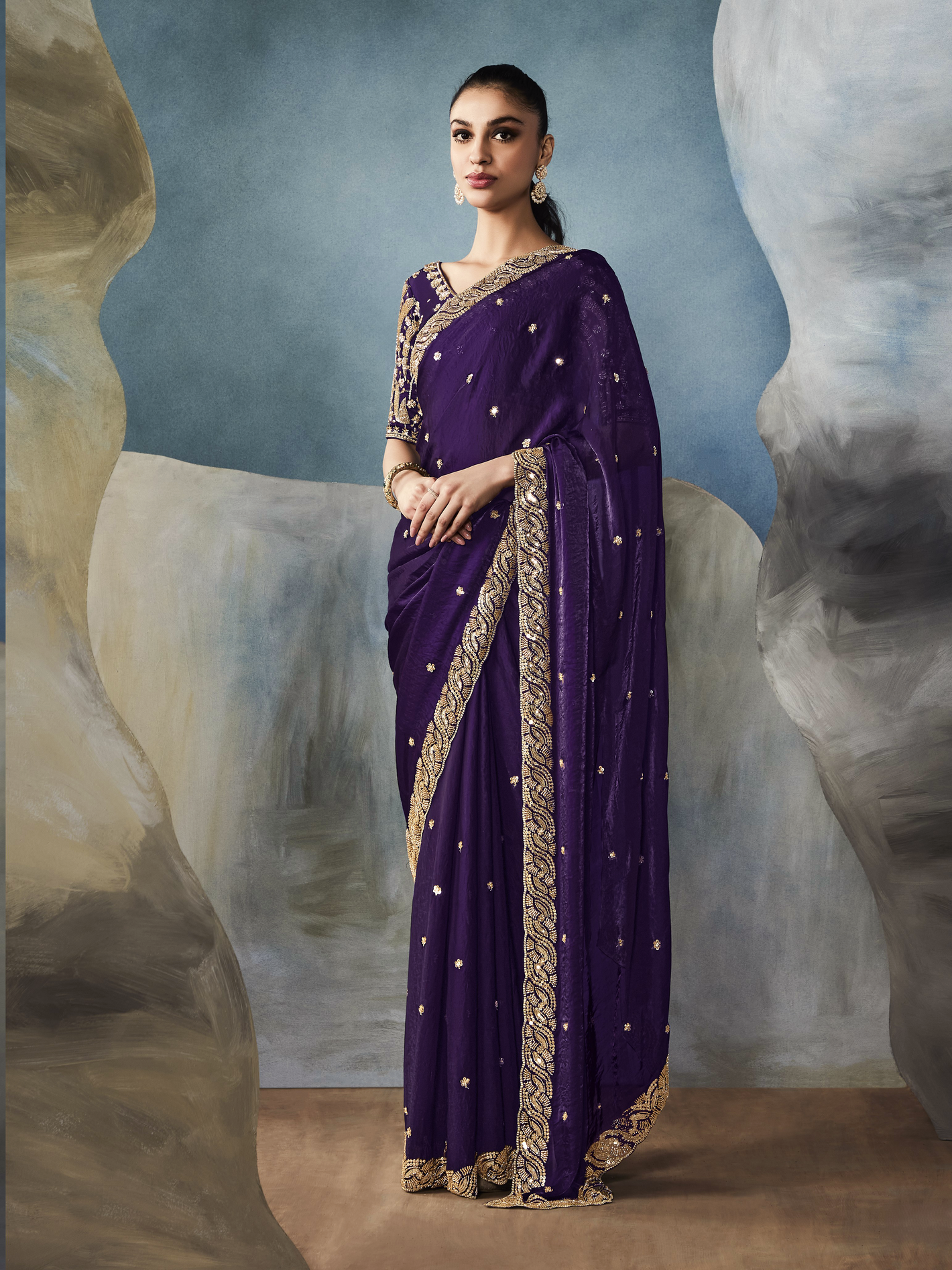 Woman wearing a purple saree with gold border against an abstract background