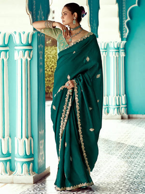 Woman in a green saree with gold details standing against a decorative blue and white wall.