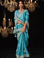Woman in a blue saree with silver patterns standing in front of chandeliers on a black background