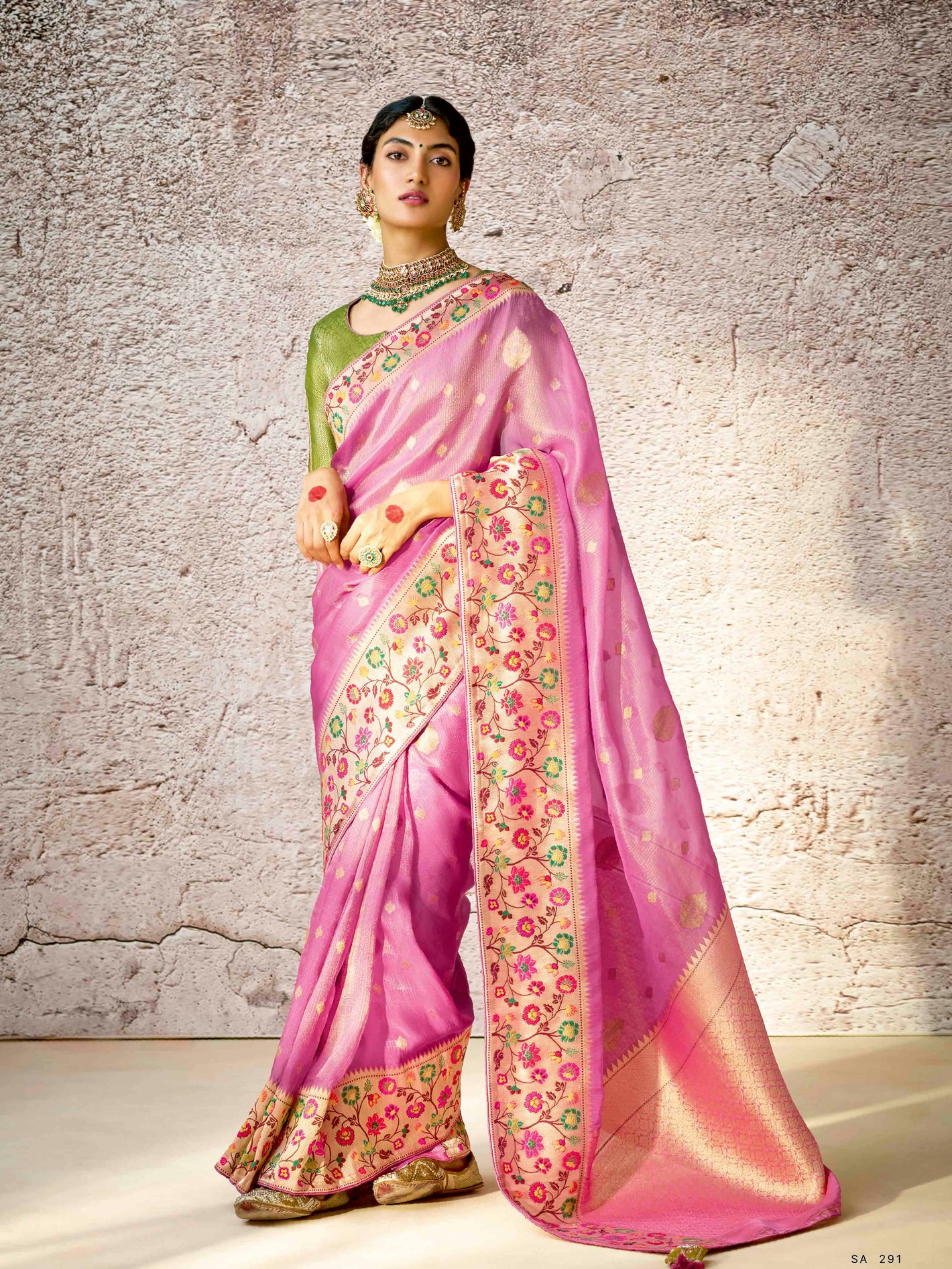 Woman wearing a pink saree with floral patterns against a textured wall.